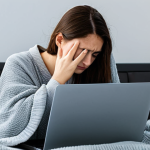 A professional, focused person, fully clothed in modest, appropriate attire, sits at a desk in a home office. Their modern laptop is clearly overheating, with visible heat subtly shimmering from the vents and an unresponsive screen. Dust is visible around the laptop's vents, which are partially blocked by a soft blanket it rests upon. The person's expression shows mild frustration or concern. The scene is well-lit, emphasizing the heat and the laptop's struggle. safe for work, appropriate content, fully clothed, professional image, perfect anatomy, correct proportions, natural pose, well-formed hands, proper finger count, natural body proportions, high quality, detailed, realistic.
